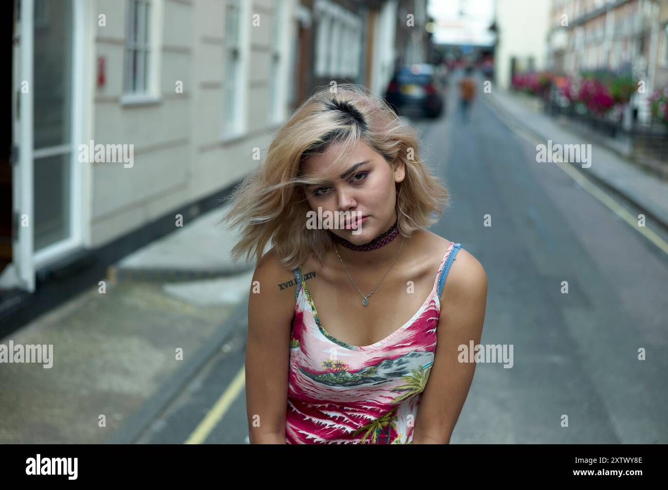 Una giovane donna bionda e un tatuaggio sul braccio si trova in una stretta strada della città, dando un look premuroso. Foto Stock