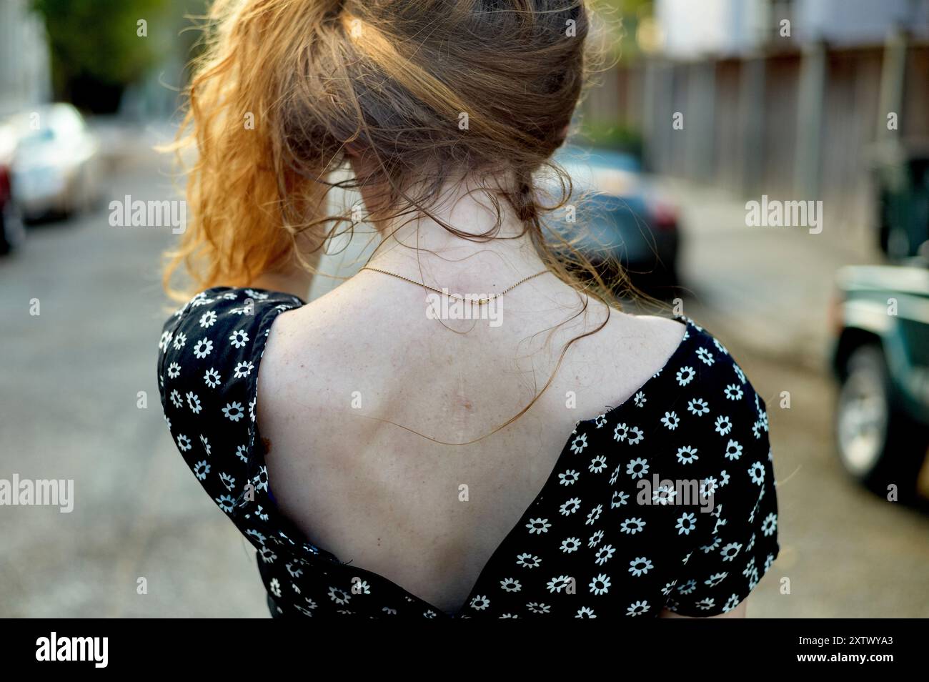 Una donna con i capelli ricci rossi che indossa un abito nero con fiori bianchi sta con la schiena alla fotocamera in una strada della città. Foto Stock