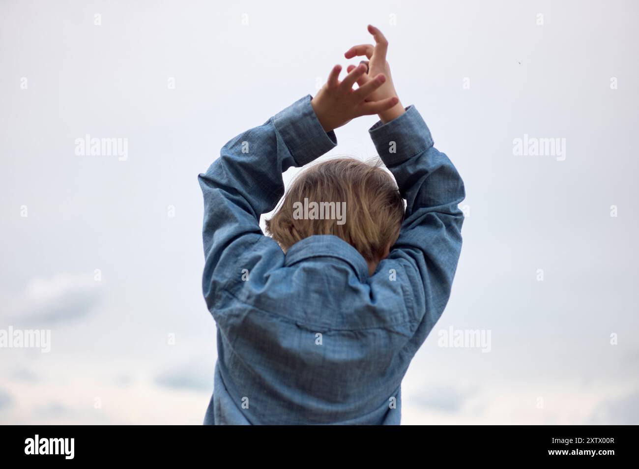 Il bambino con la camicia in denim raggiunge il cielo con le braccia allungate, appoggiandosi su uno sfondo morbido e nuvoloso. Foto Stock