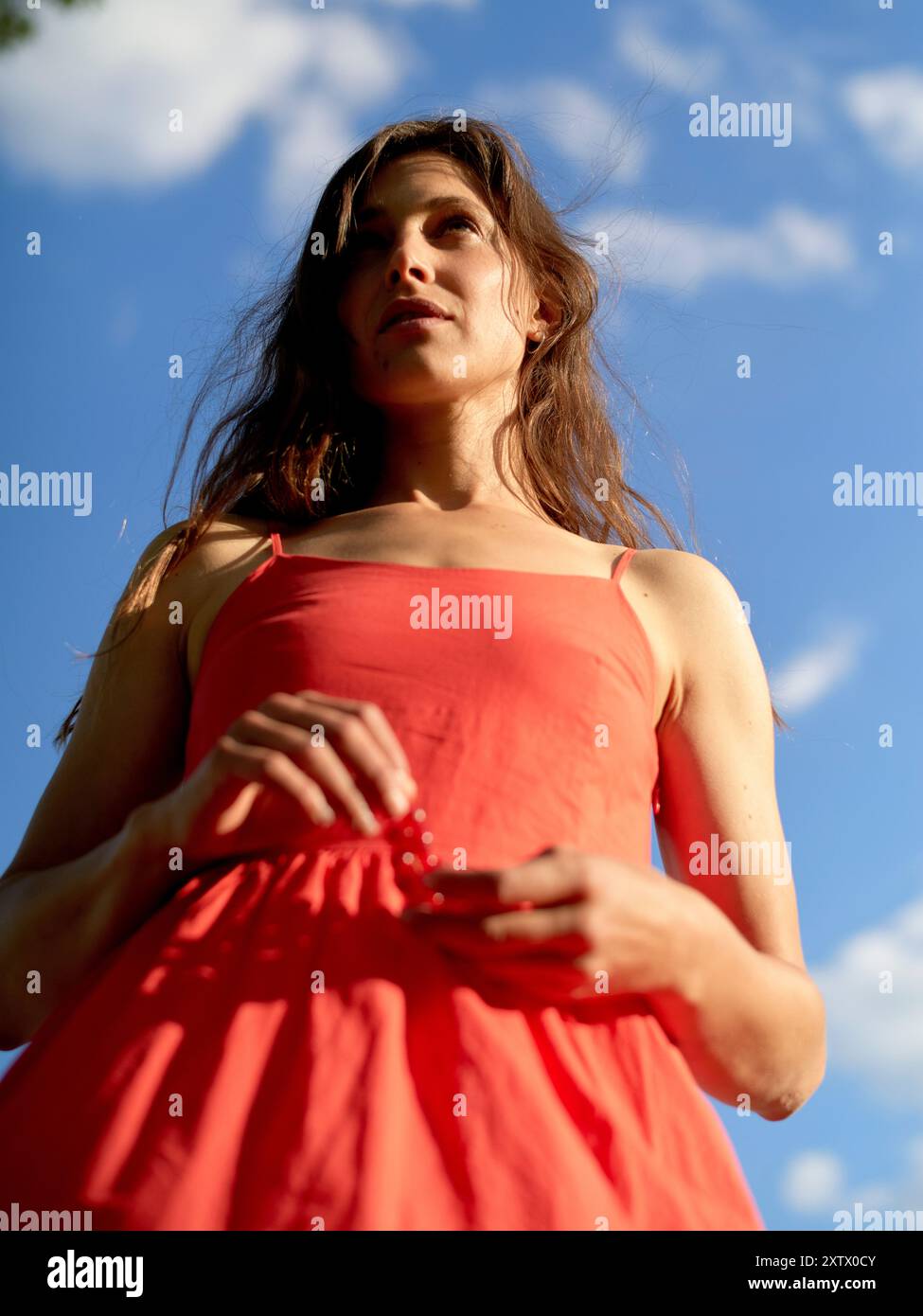 Una donna con un abito arancione si erge contro un cielo blu con nuvole bianche. Foto Stock