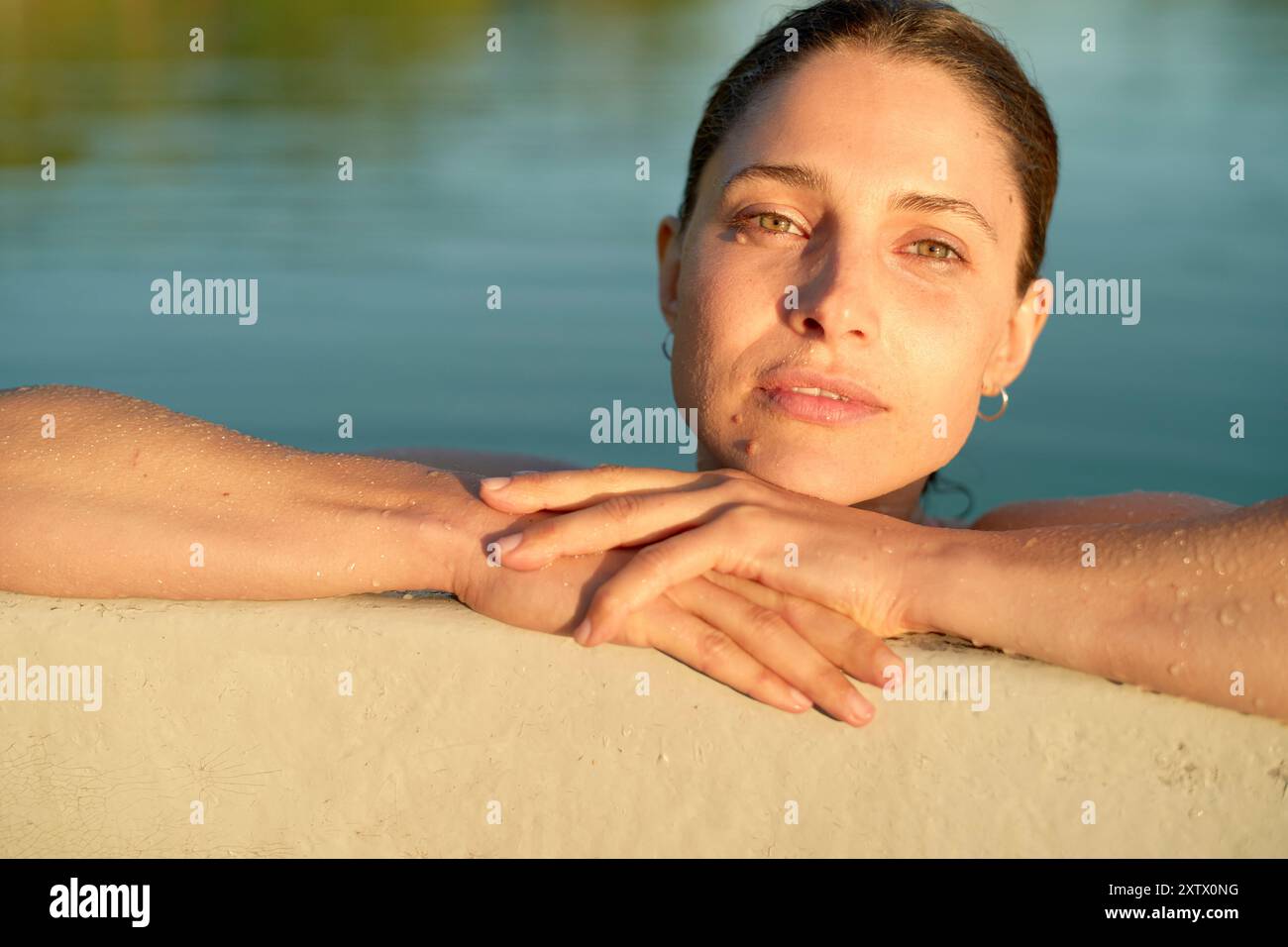 Donna serena con pelle bagnata che riposa sul bordo di una piscina durante il tramonto. Foto Stock