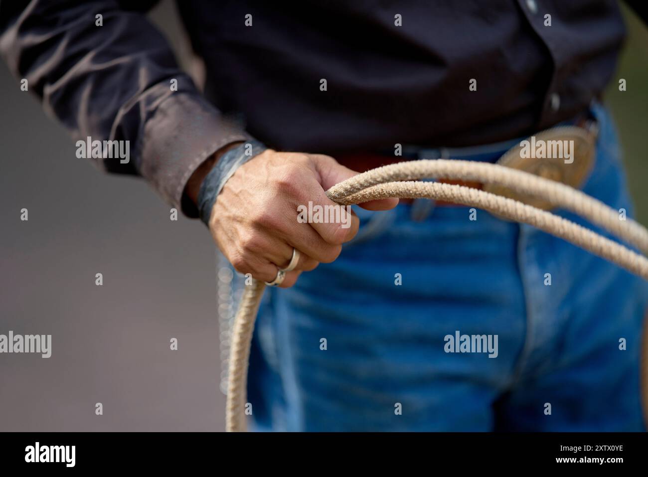 Primo piano delle mani di una persona che avvolge una corda, vestita con jeans e una camicia scura a maniche lunghe, con sfondo sfocato. Foto Stock