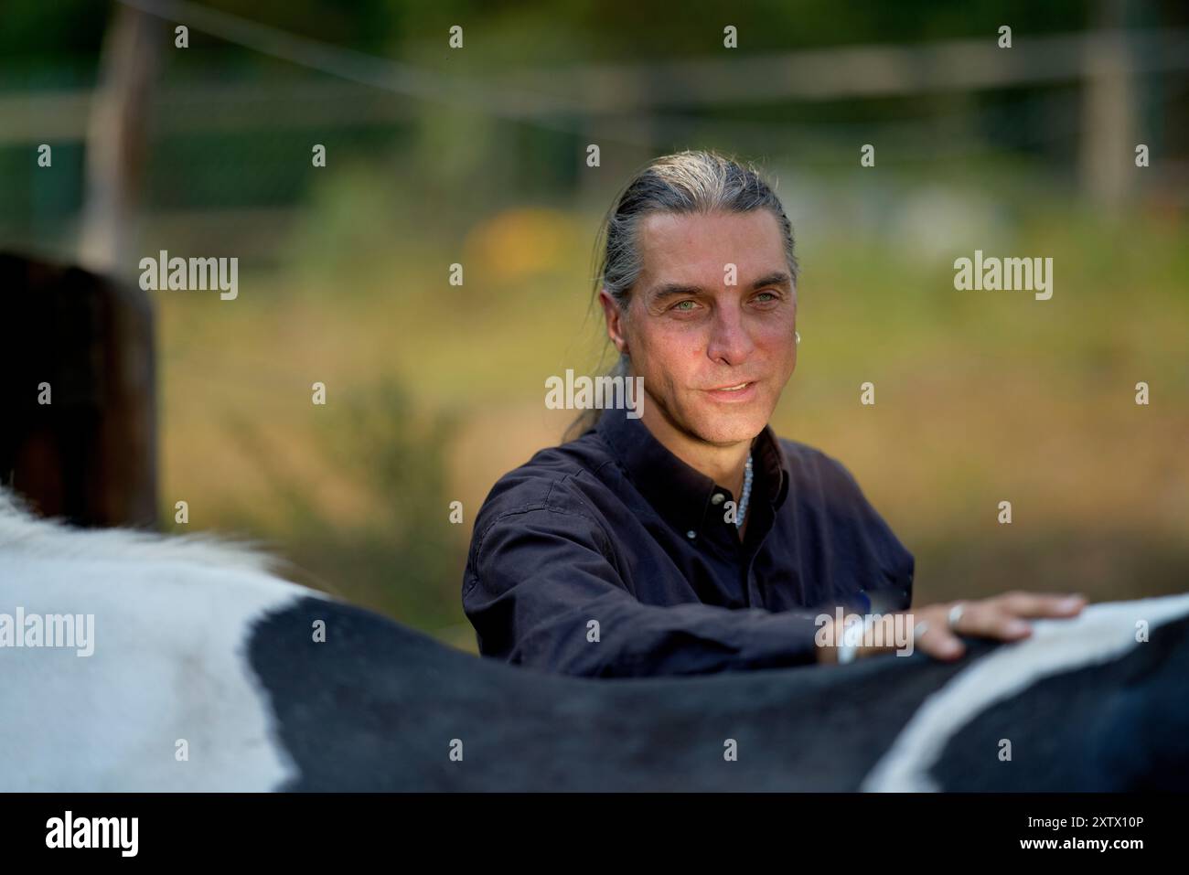 Donna sorridente con lunghi capelli grigi che riposa il braccio sulla schiena di un cavallo in natura. Foto Stock