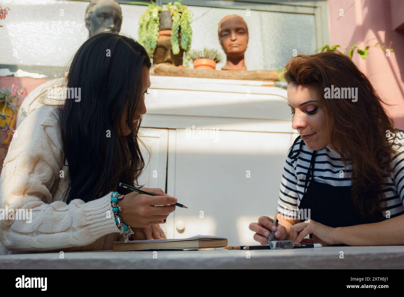 Insegnante d'arte femminile a casa che spiega le tecniche di disegno della grafite, due donne sedute a casa che praticano e studiano arte, usando quaderni e colori Foto Stock