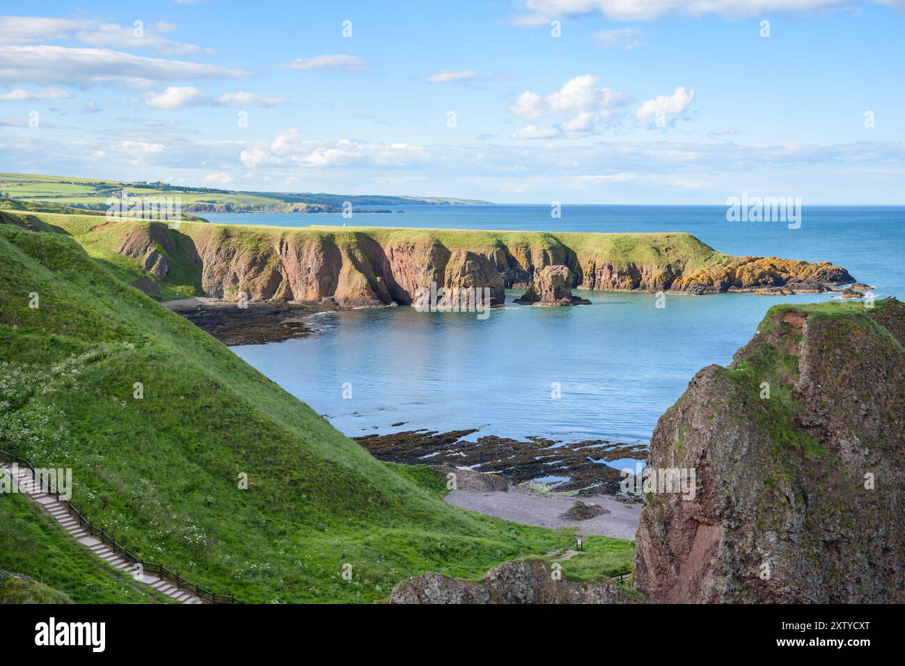 Lungo una costa frastagliata in una soleggiata giornata estiva. In primo piano c'è una scala vuota che scende da una scogliera erbosa fino a una piccola spiaggia rocciosa Foto Stock