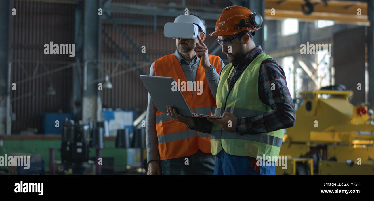 Due tecnici professionisti del settore che indossano uniforme protettiva e caschi si trovano presso lo stabilimento di produzione. Un tecnico maturo utilizza un visore VR, un dipendente adulto lavora su un computer portatile. Al rallentatore. Foto Stock