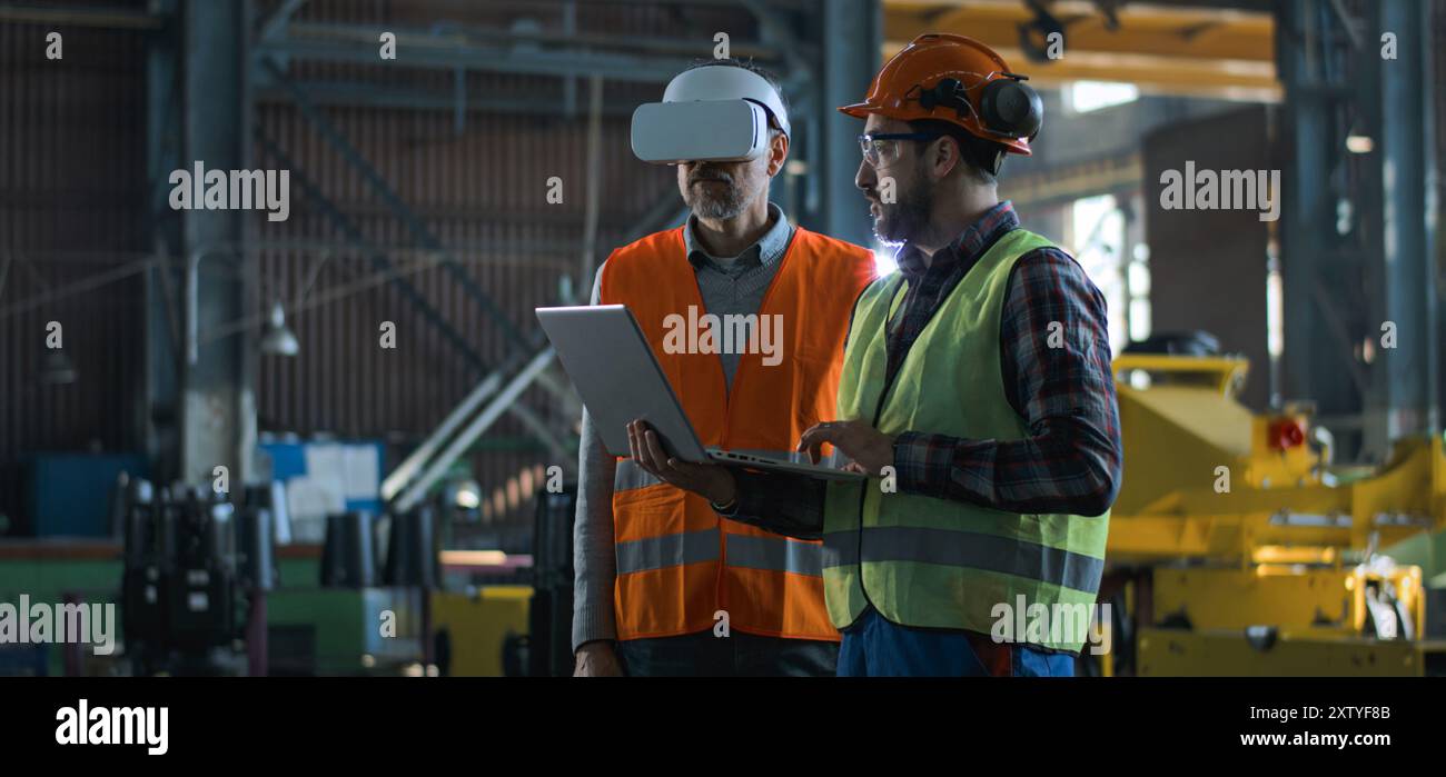 Due tecnici professionisti del settore che indossano uniforme protettiva e caschi si trovano presso lo stabilimento di produzione. Un tecnico maturo utilizza un visore VR, un dipendente adulto lavora su un computer portatile. Al rallentatore. Foto Stock