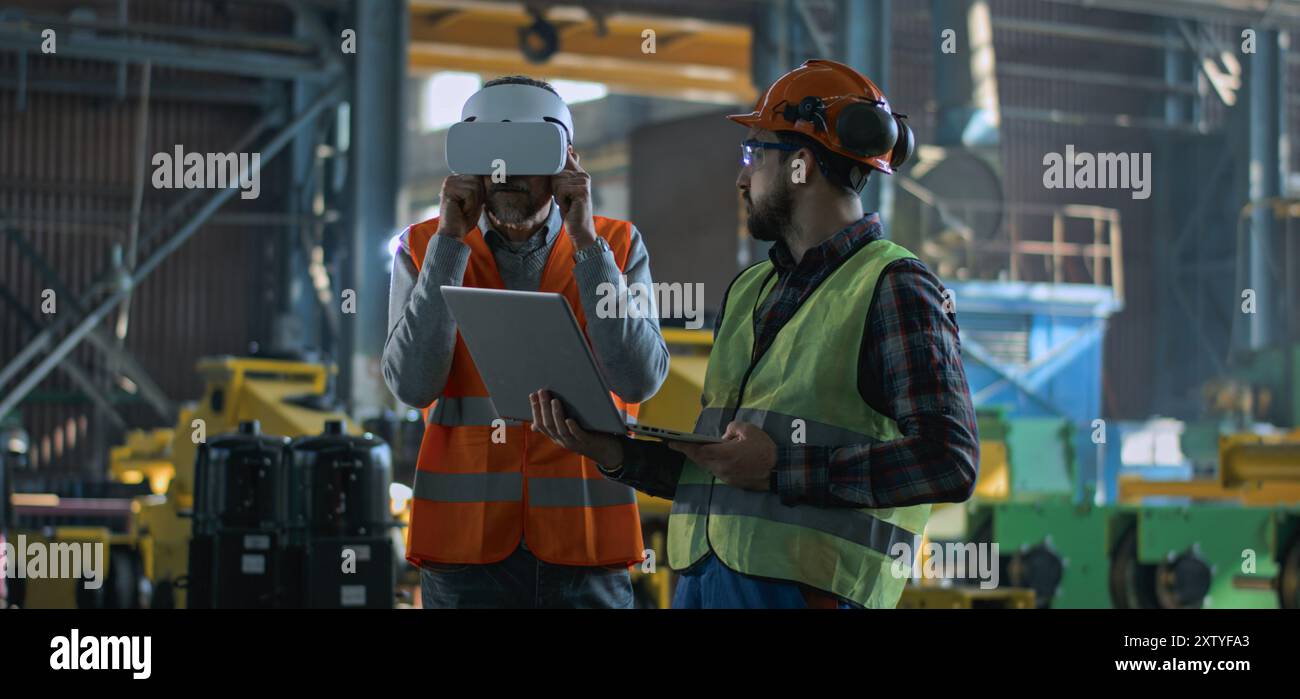 Due tecnici professionisti del settore che indossano uniforme protettiva e caschi si trovano presso lo stabilimento di produzione. Un tecnico maturo utilizza un visore VR, un dipendente adulto lavora su un computer portatile. Al rallentatore. Foto Stock