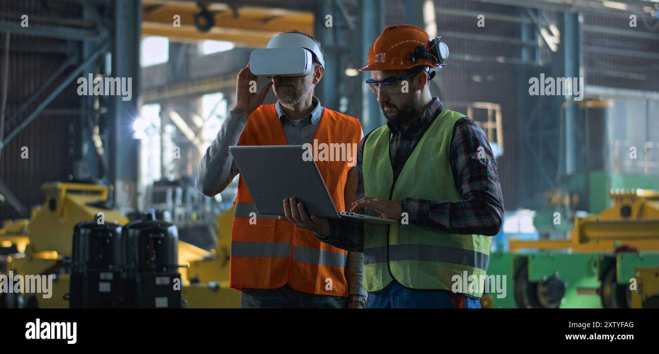 Due tecnici professionisti del settore che indossano uniforme protettiva e caschi si trovano presso lo stabilimento di produzione. Un tecnico maturo utilizza un visore VR, un dipendente adulto lavora su un computer portatile. Al rallentatore. Foto Stock