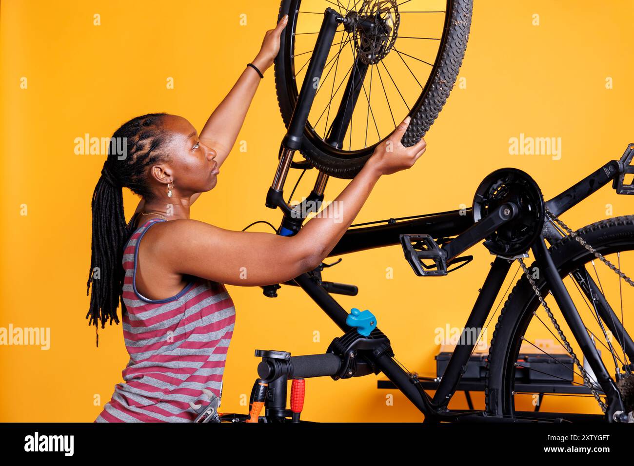 Meticolosa signora afroamericana ripara la moto rotta, regolando gli ingranaggi e fissando la ruota con precisione esperta. Donna nera sportiva che esamina e cambia pneumatico della bicicletta. Foto Stock