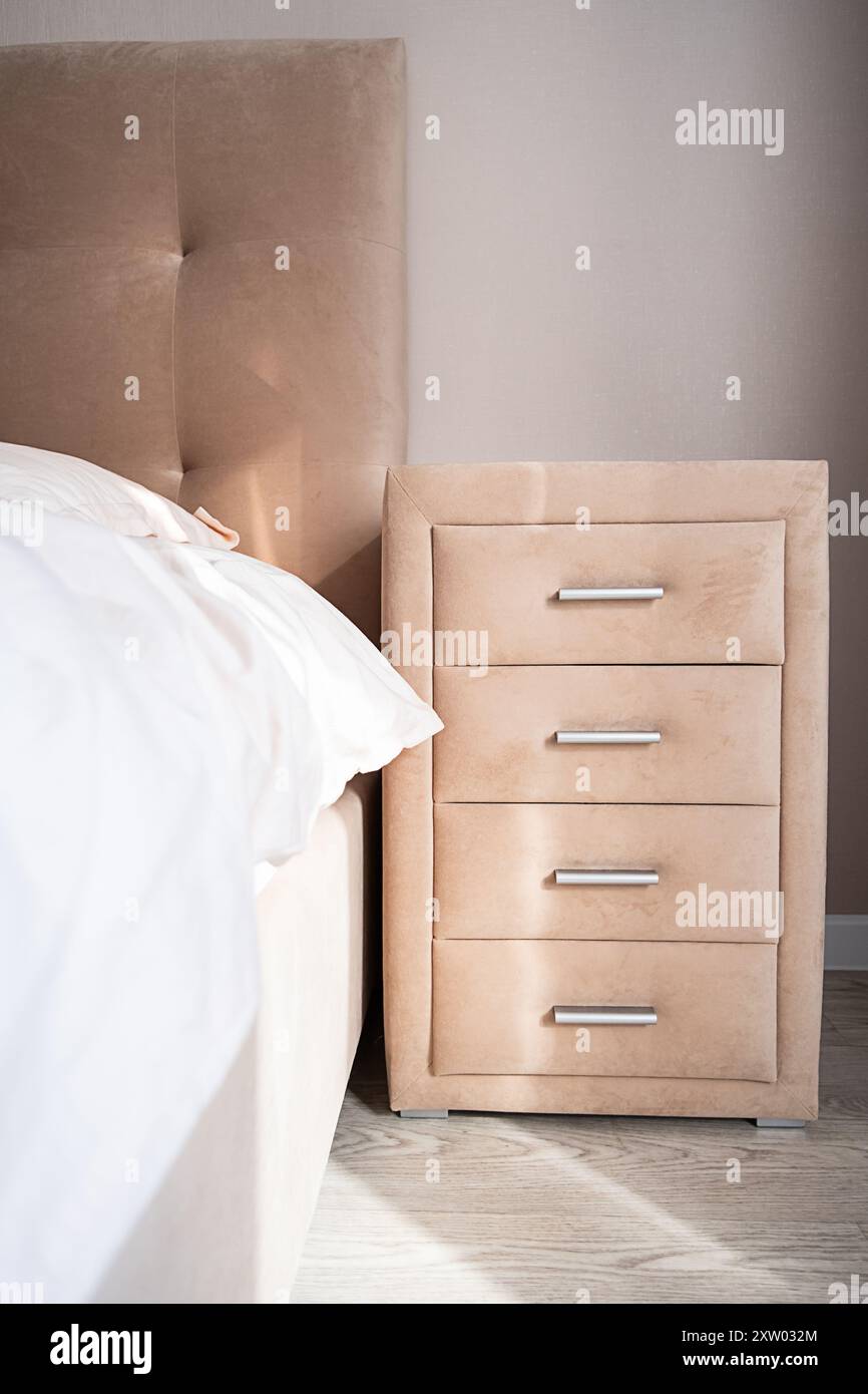 Comò beige con quattro cassetti vicino al letto. Camera ben organizzata con luce naturale. Foto Stock