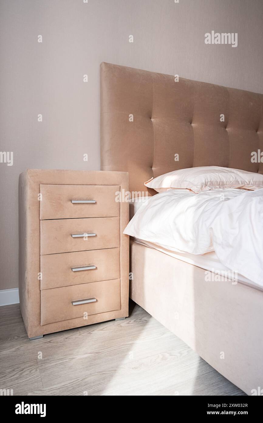 Comò beige con quattro cassetti vicino al letto. Camera ben organizzata con luce naturale. Foto Stock