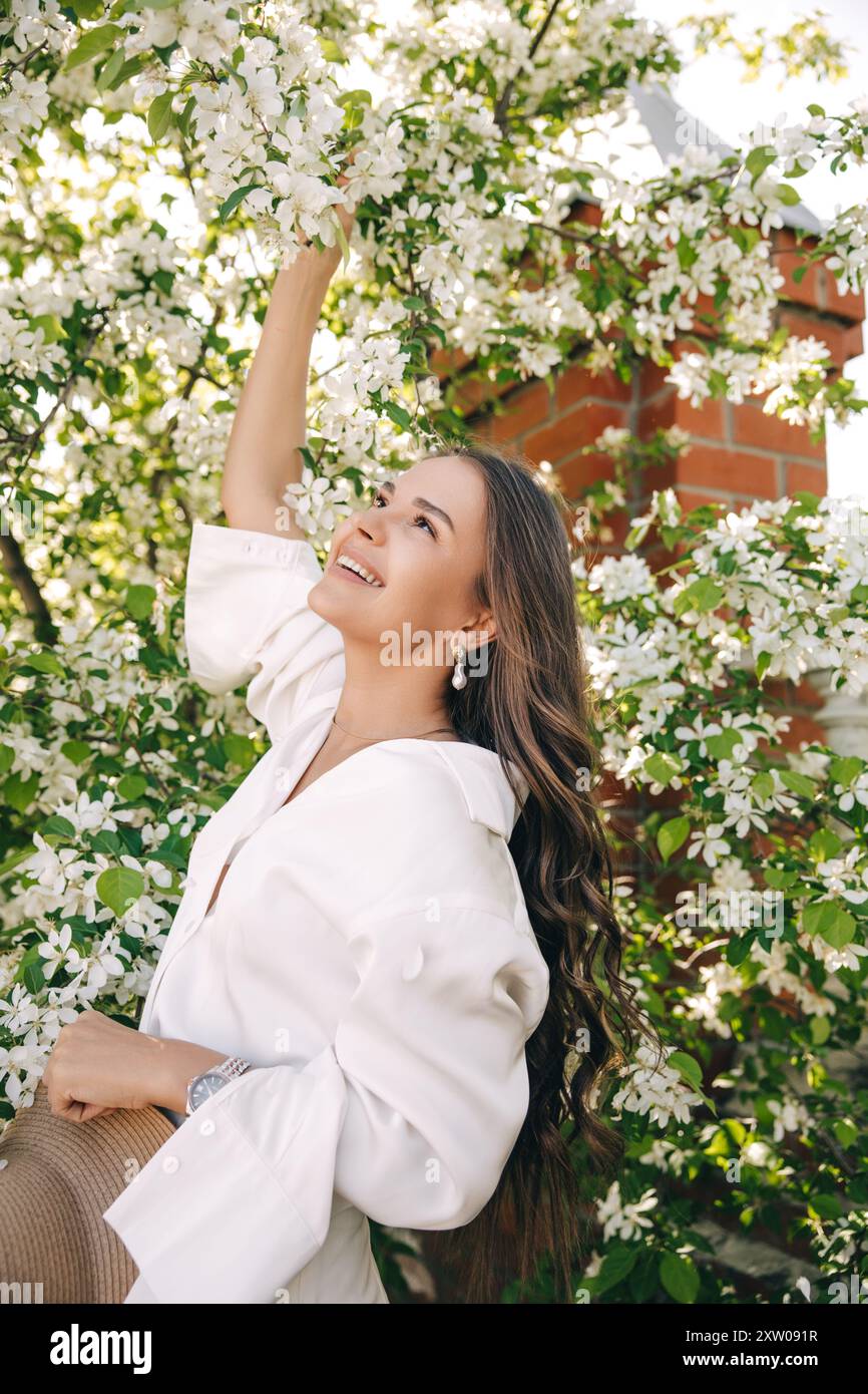 Ritratto di una bella ragazza con i capelli lunghi vicino a un albero in fiore in abiti bianchi alla moda Foto Stock