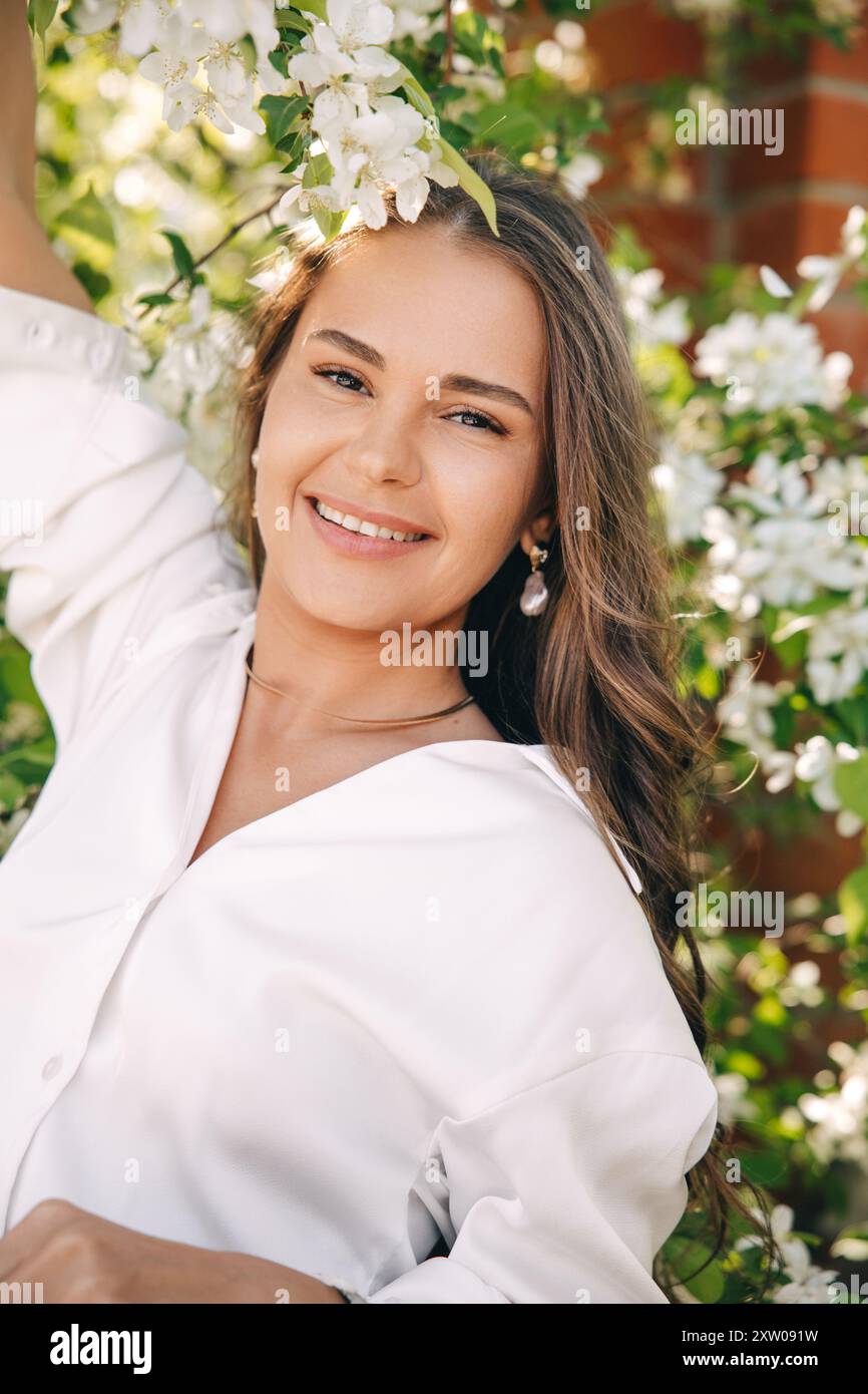 Ritratto di una bella ragazza con i capelli lunghi vicino a un albero in fiore in abiti bianchi alla moda Foto Stock