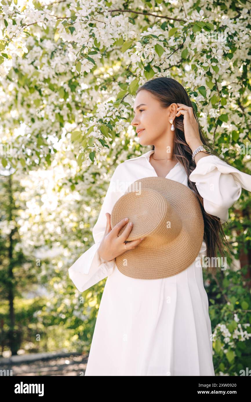 Ritratto di una bella ragazza con i capelli lunghi vicino a un albero in fiore in abiti bianchi alla moda Foto Stock