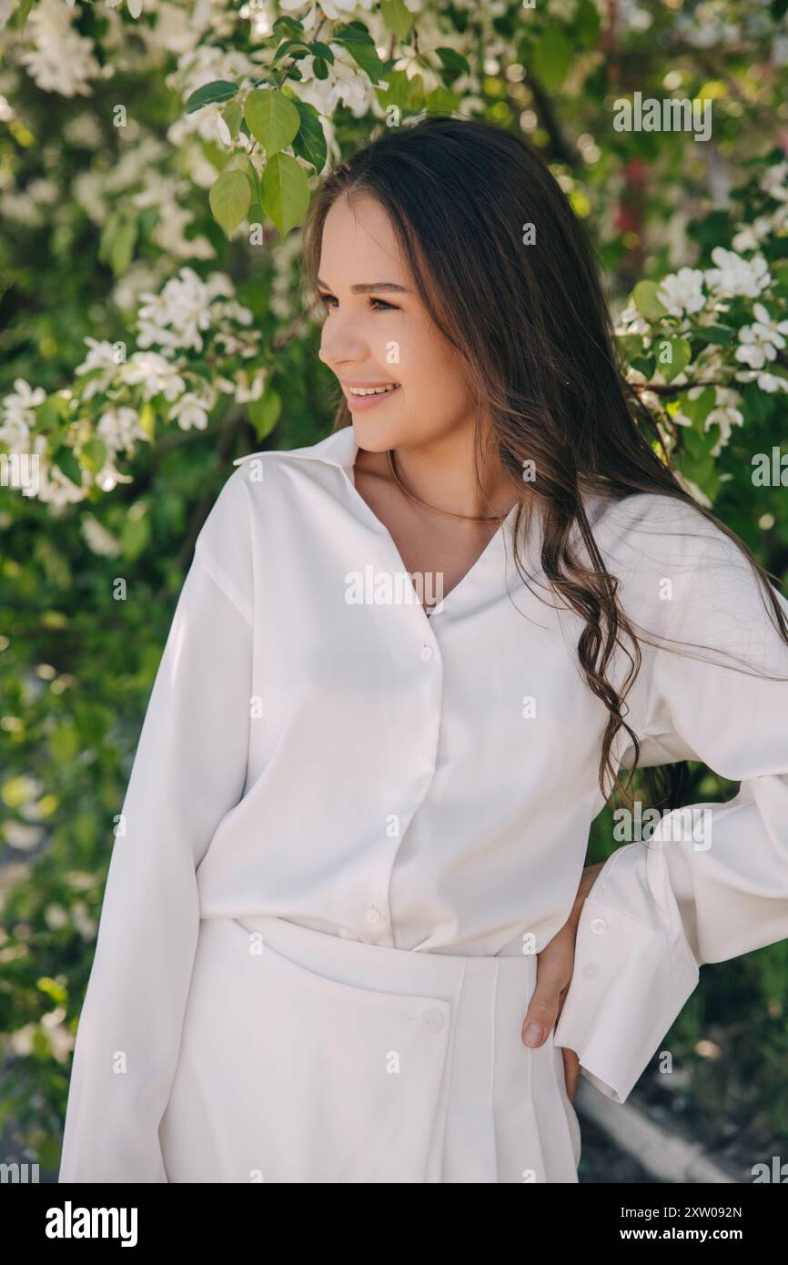 Ritratto di una bella ragazza con i capelli lunghi vicino a un albero in fiore in abiti bianchi alla moda Foto Stock