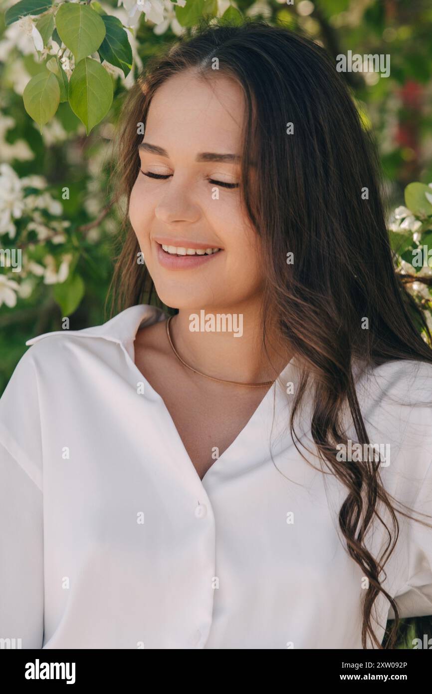 Ritratto di una bella ragazza con i capelli lunghi vicino a un albero in fiore in abiti bianchi alla moda Foto Stock