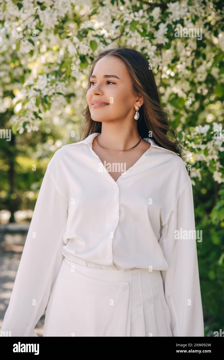 Ritratto di una bella ragazza con i capelli lunghi vicino a un albero in fiore in abiti bianchi alla moda Foto Stock