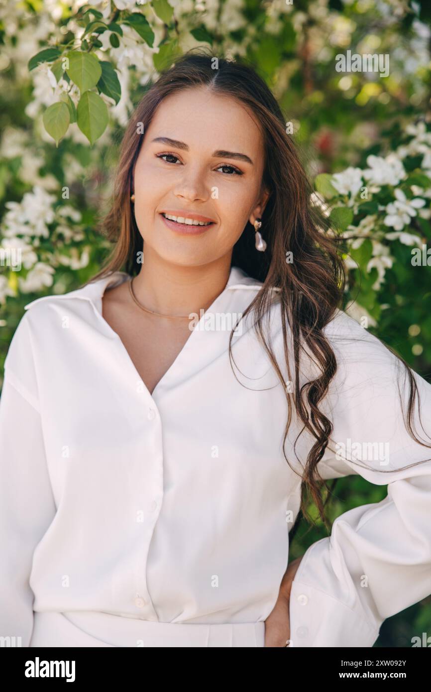 Ritratto di una bella ragazza con i capelli lunghi vicino a un albero in fiore in abiti bianchi alla moda Foto Stock