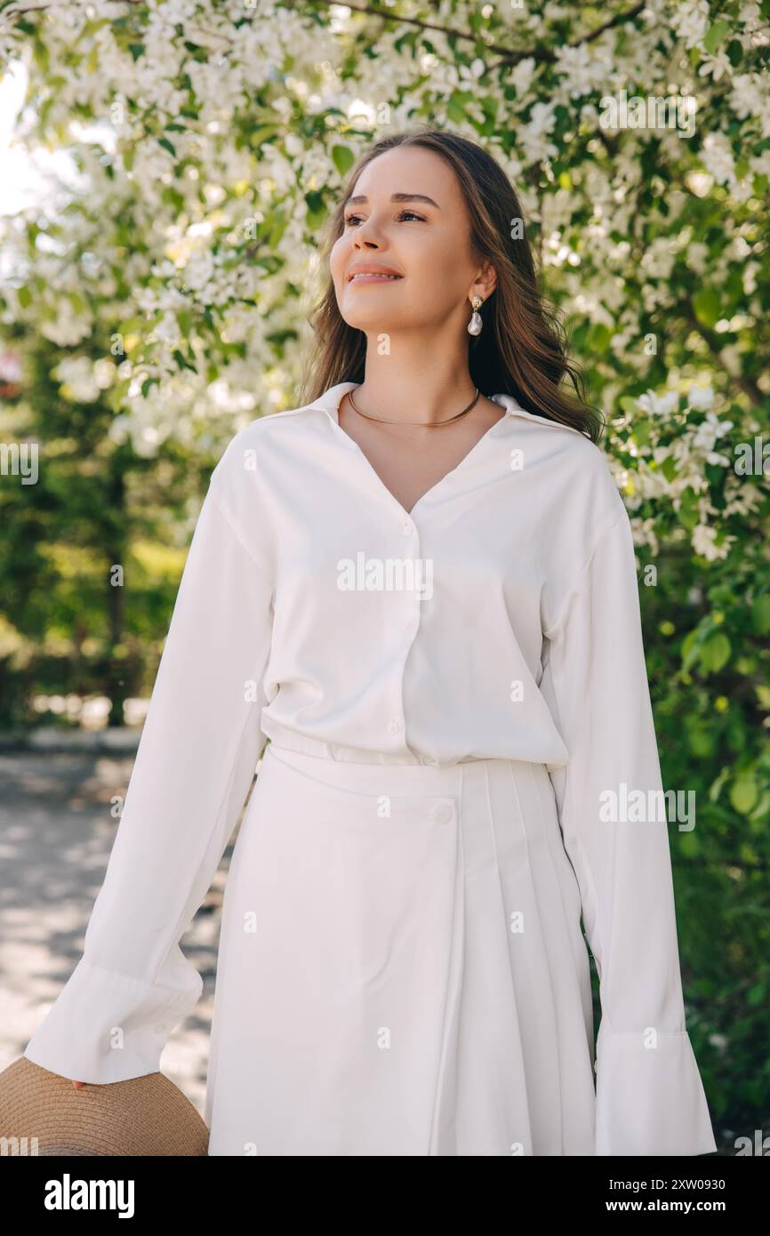 Ritratto di una bella ragazza con i capelli lunghi vicino a un albero in fiore in abiti bianchi alla moda Foto Stock