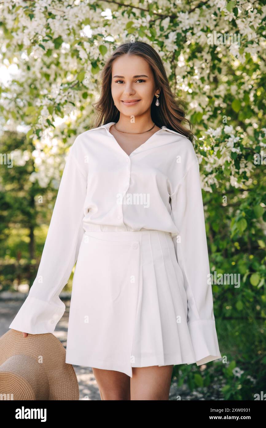 Ritratto di una bella ragazza con i capelli lunghi vicino a un albero in fiore in abiti bianchi alla moda Foto Stock