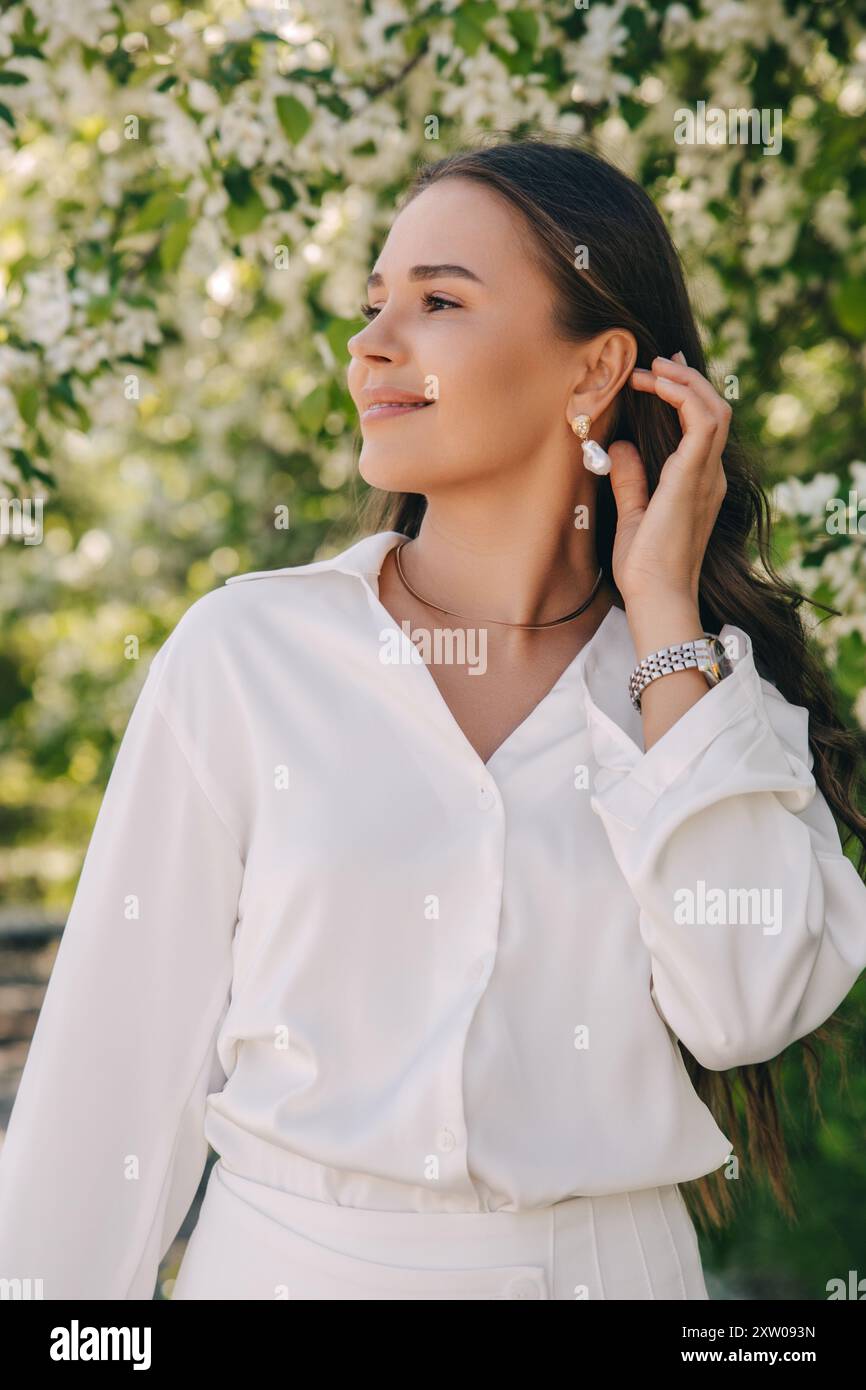 Ritratto di una bella ragazza con i capelli lunghi vicino a un albero in fiore in abiti bianchi alla moda Foto Stock
