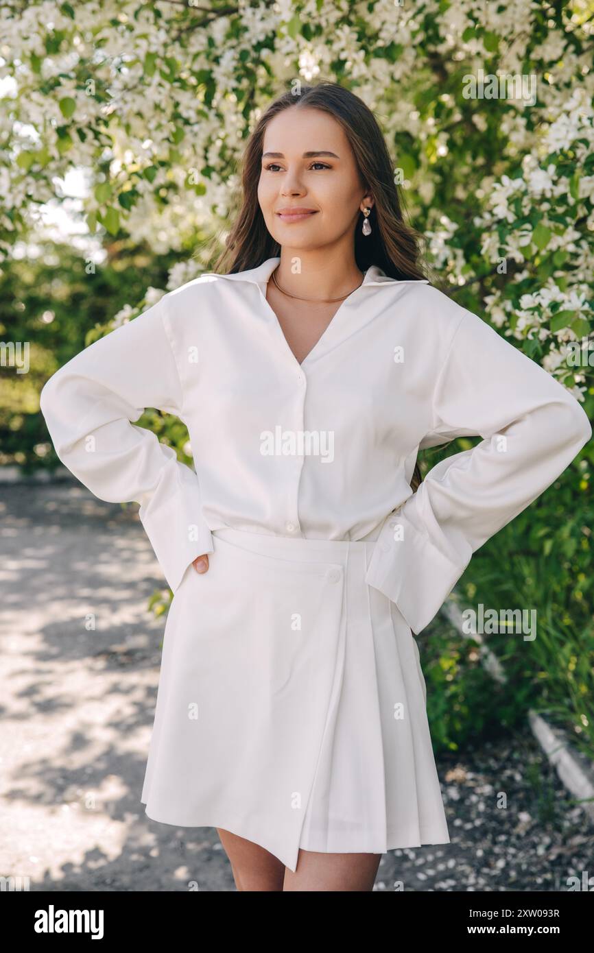 Ritratto di una bella ragazza con i capelli lunghi vicino a un albero in fiore in abiti bianchi alla moda Foto Stock