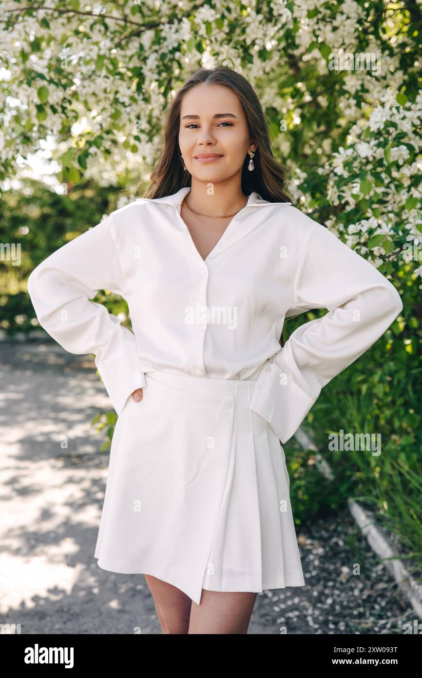 Ritratto di una bella ragazza con i capelli lunghi vicino a un albero in fiore in abiti bianchi alla moda Foto Stock