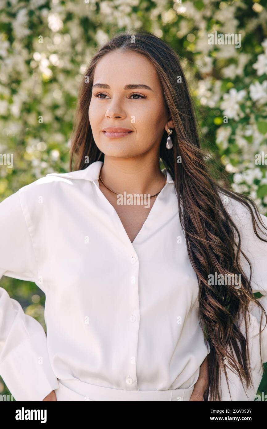 Ritratto di una bella ragazza con i capelli lunghi vicino a un albero in fiore in abiti bianchi alla moda Foto Stock