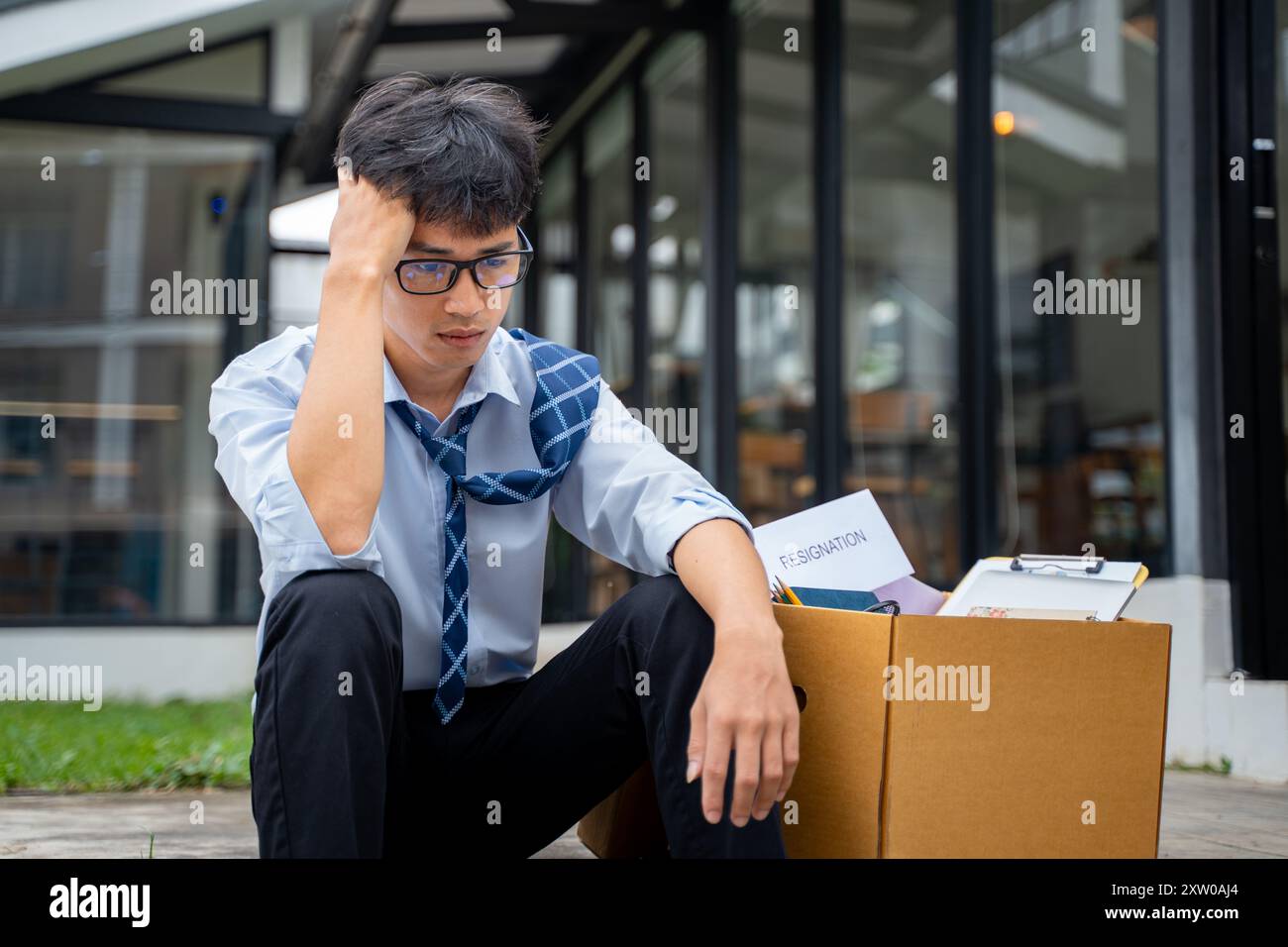 Triste giovane uomo d'affari seduto su una strada laterale curva con una scatola piena di cose del suo ufficio accanto a lui Foto Stock