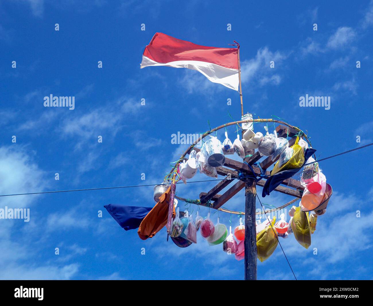 In cima a un palo sono appesi vari premi utilizzati per una gara di panjat pinang. Sopra di loro, la bandiera rossa e bianca sventola contro un cielo blu brillante. Foto Stock