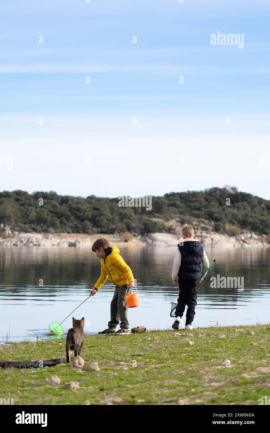 Due bambini giocano a pesca in un lago in compagnia del loro gatto Foto Stock