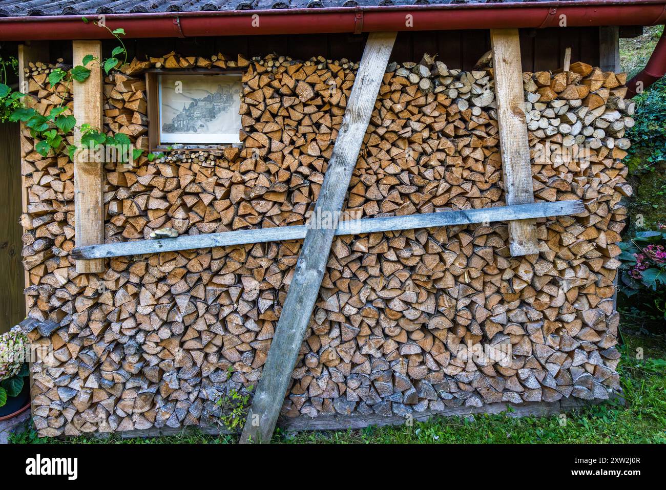Una catasta di legna sul Bruderweg a Berneck, Altensteig, Baden-Württemberg, Germania, si apre su una vista attraverso una finestra ritagliata. Due travi diagonali sostengono la pila contro l'edificio vicino. Foto Stock