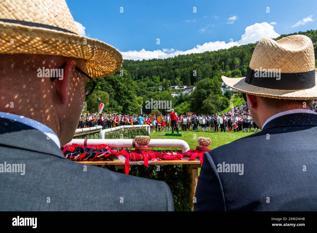 Le corone dei pastori, il cosiddetto "Schäppel", sono pronte su un tavolo in primo piano, per essere posate sui vincitori dopo la corsa dei pastori alla Schäferlauf 2024 a Wildberg, Baden-Württemberg, Germania. Due spettatori con cappelli di paglia osservano un concerto di banda sulla piazza del festival nella prateria oltre. L'evento fa parte della Schäferlauf, riconosciuta come patrimonio culturale immateriale dall'UNESCO dal 2018 – 300 anni di tradizione dal 1723. Foto Stock