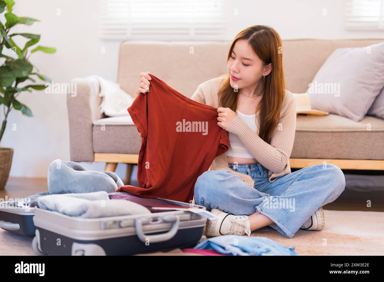 Una giovane donna è seduta sul pavimento, a controllare gli articoli su una lista di imballaggio mentre organizza la sua valigia per un viaggio, assicurandosi che tutto sia in ordine. Foto Stock