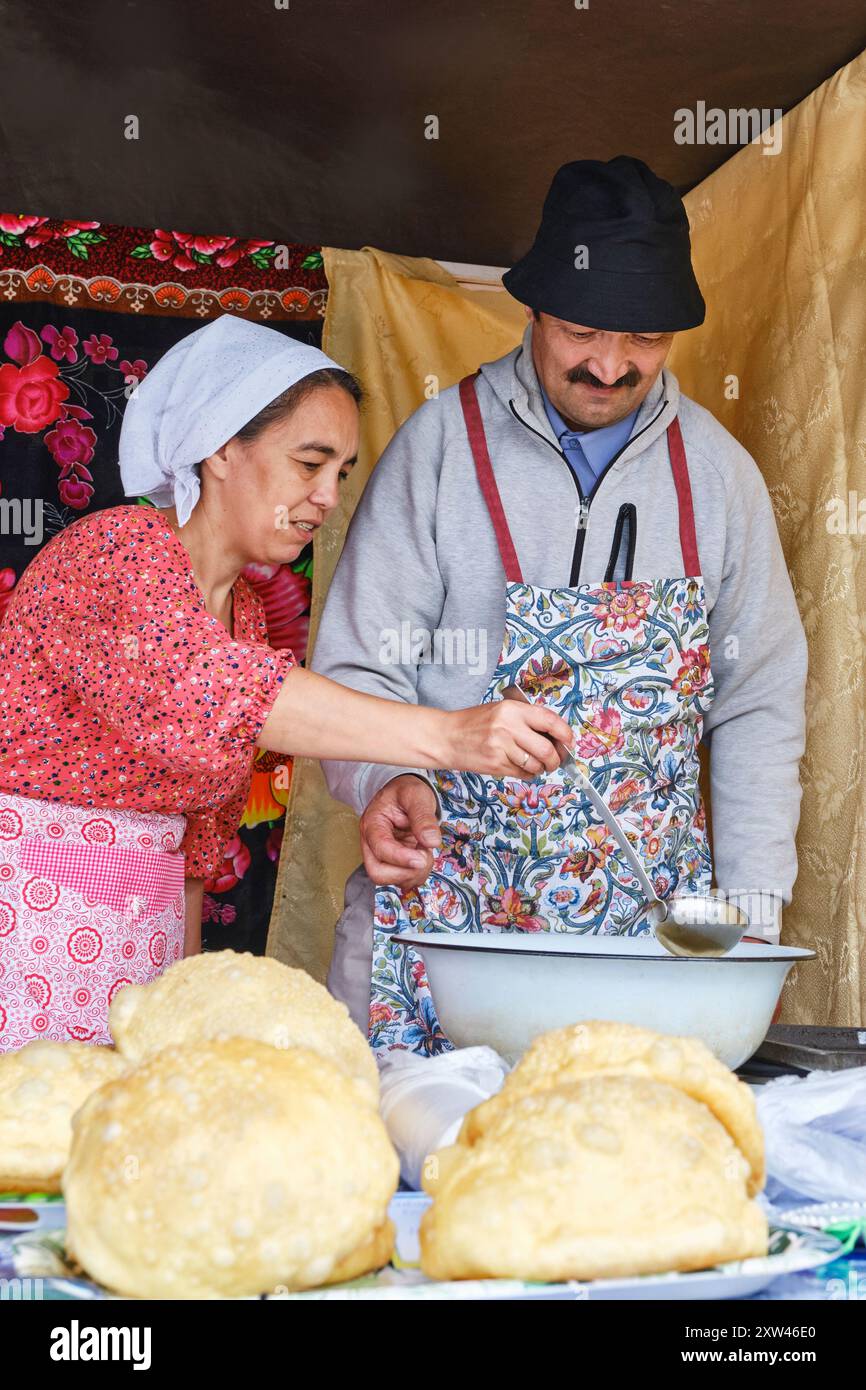 Tobolsk, Russia - 20 luglio 2024: La cuoca donna asiatica mostra all'uomo anziano come friggere il boortsog, tenendo il mestolo. Master class culinaria sulla cottura al forno. Tataro Foto Stock