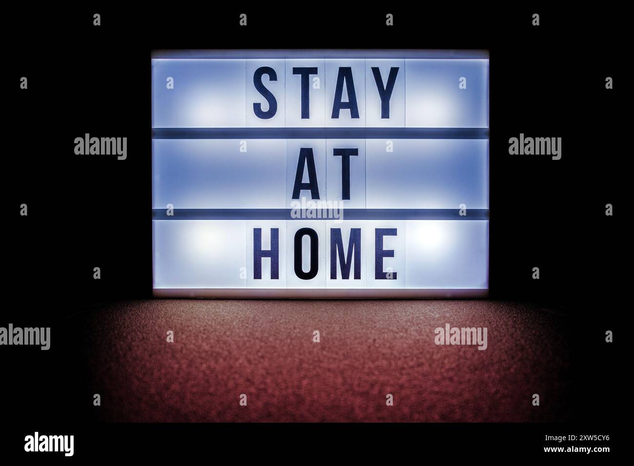 Una scatola luminosa che visualizza il messaggio rimane a casa in grassetto, lettere nere, illuminata su uno sfondo scuro. Foto Stock