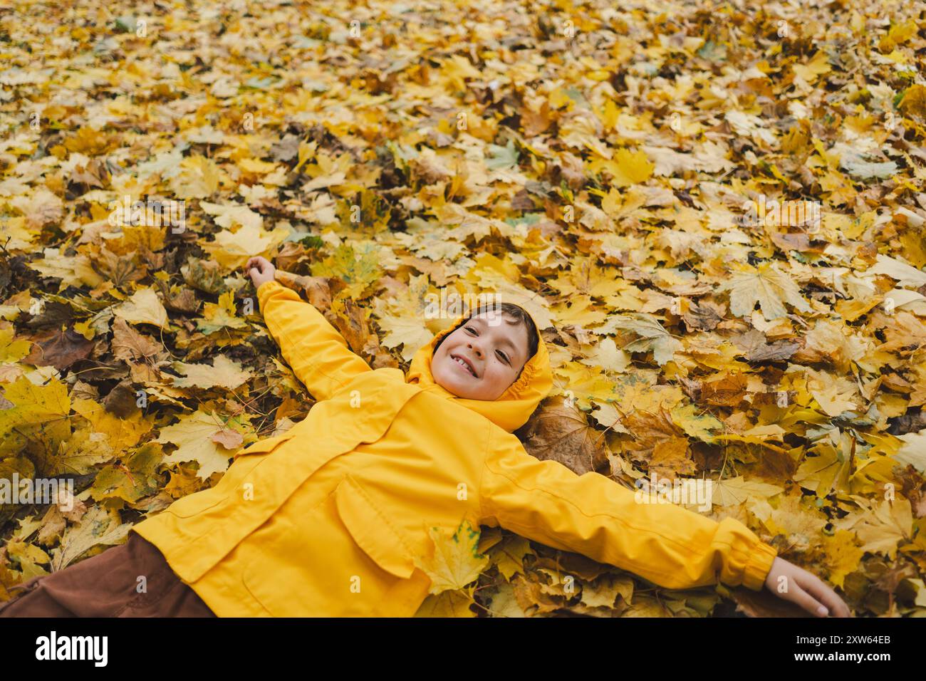 Gioioso bambino in impermeabile giallo giacente in foglie autunnali in un parco Foto Stock