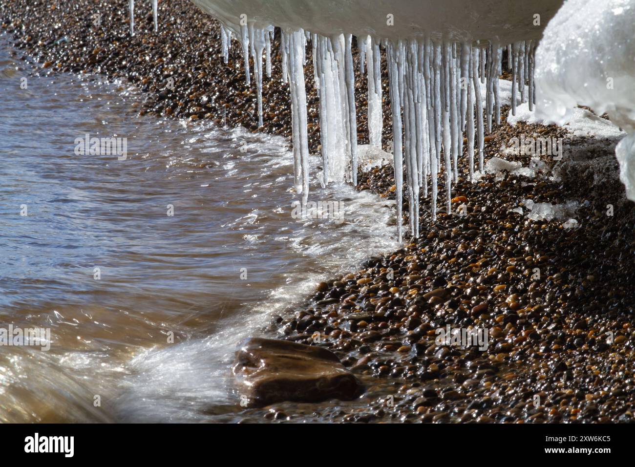 Da un fondo di ghiaccio adagiato su una spiaggia di ciottoli, molti ghiaccioli sono appesi. Le onde rotolano sulla riva. Foto Stock