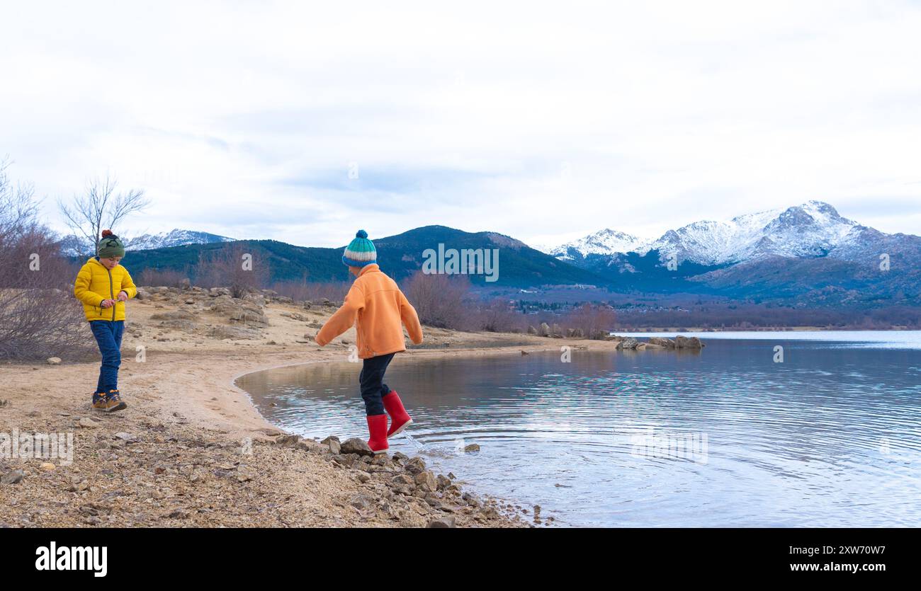 Due bambini che giocano sulla riva di un lago in una giornata fredda con montagne innevate sullo sfondo Foto Stock