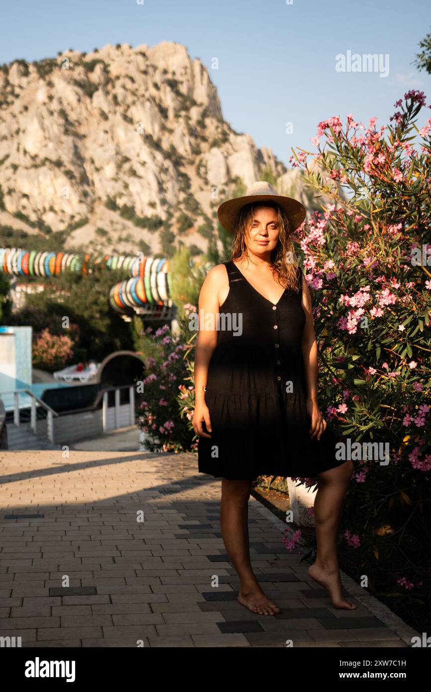 Woman Hat Mountain Landscape - Una donna che indossa un cappello si trova di fronte a una montagna e a un cespuglio. Foto Stock