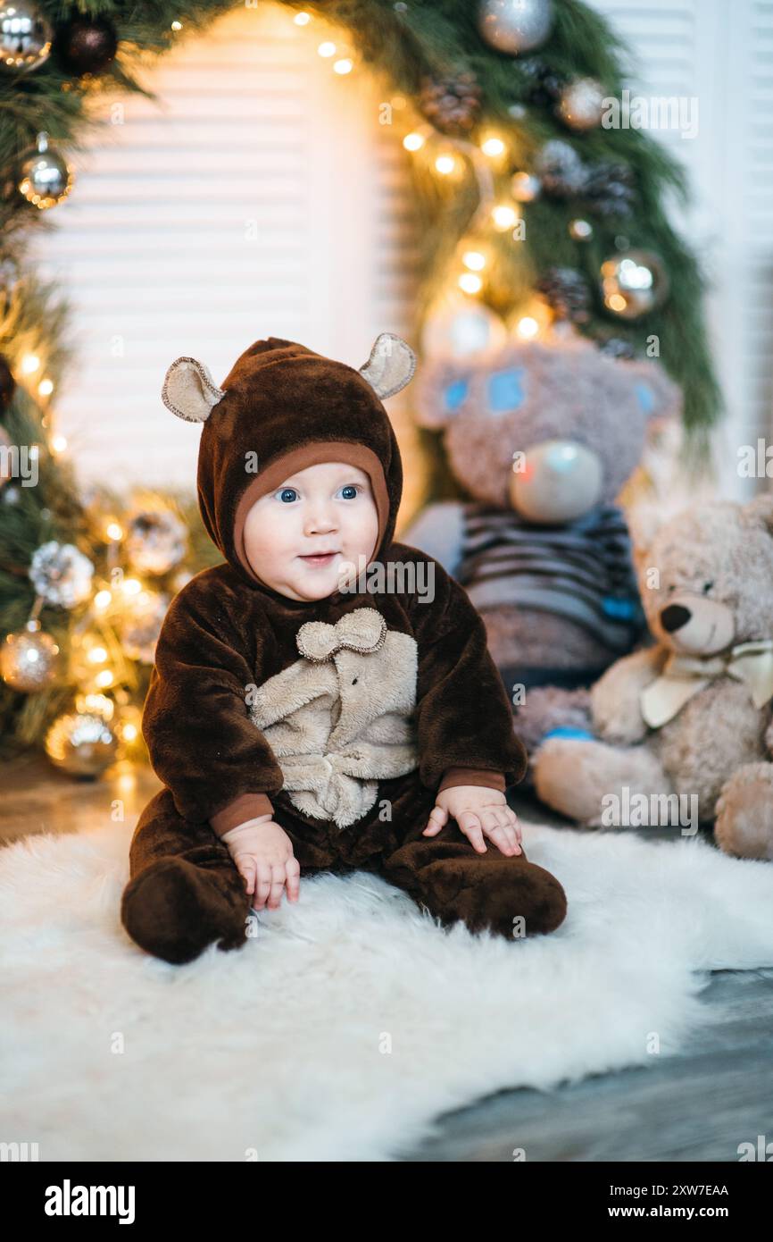 Un bambino piccolo in un costume marrone orsacchiotto siede sul pavimento vicino all'albero di Capodanno Foto Stock