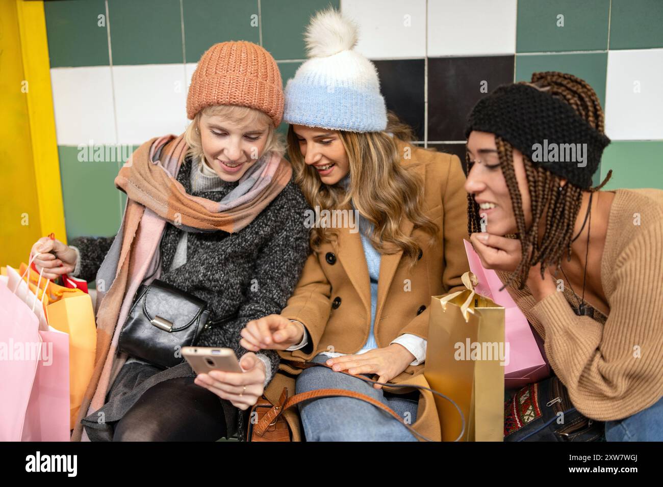 Diverse giovani amiche che condividono i contenuti dello smartphone dopo lo shopping. Indossa abiti invernali alla moda con borse colorate. Connettività e modalità digitali Foto Stock