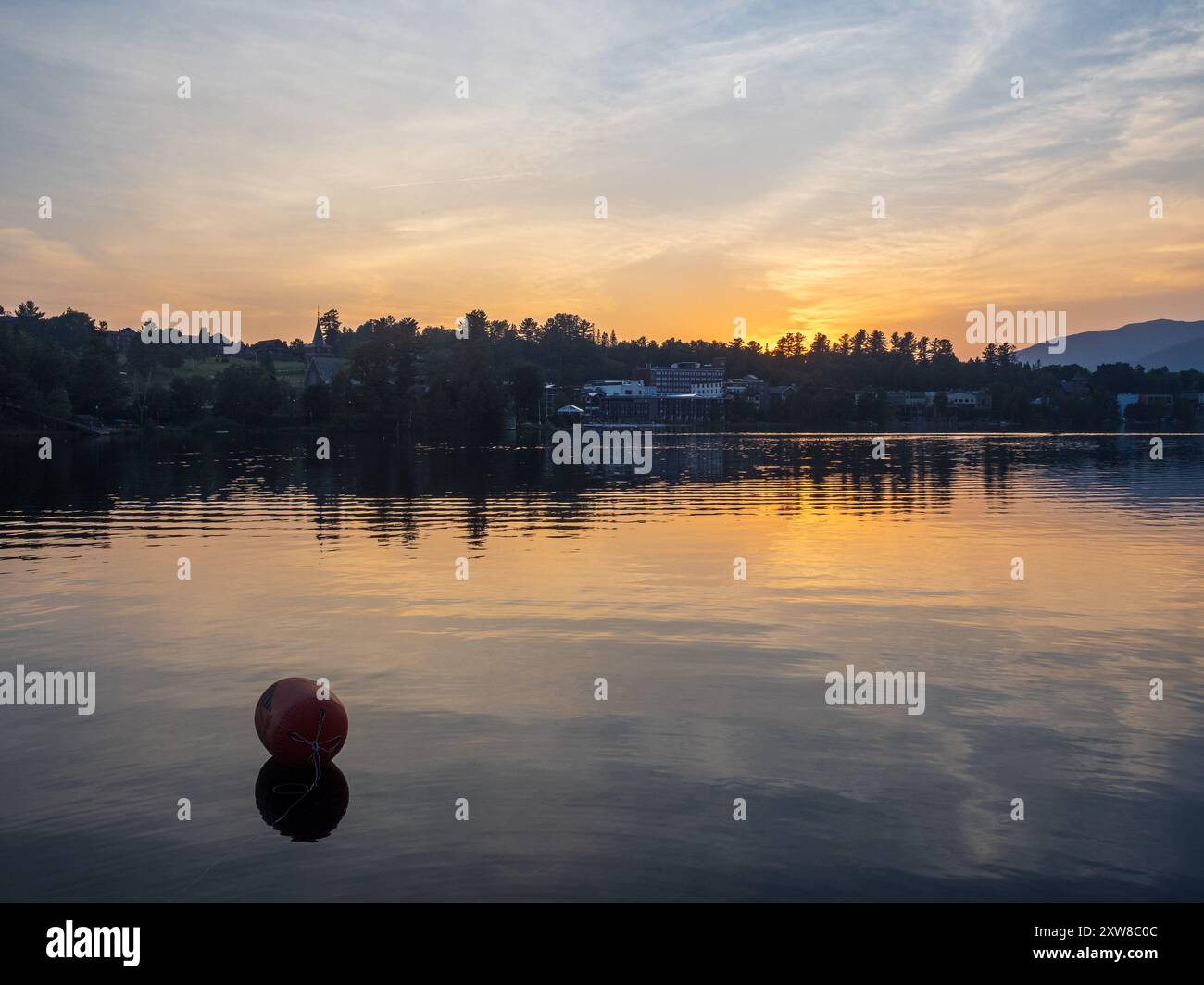 Un tranquillo tramonto dipinge il lago Placid in tonalità calde, con una boa che galleggia in primo piano e le maestose montagne Adirondack che si innalzano attraverso lo specchio L. Foto Stock