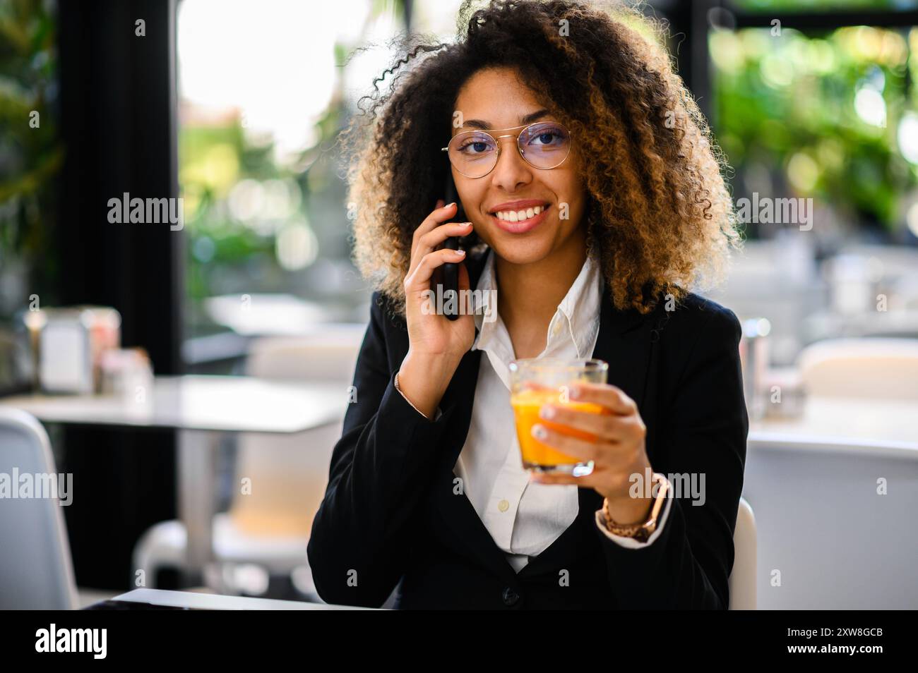 Una giovane donna sorridente in abito da lavoro che parla al telefono cellulare in un bar con un drink in mano Foto Stock