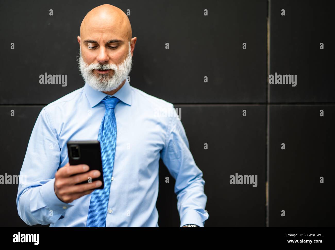 Un uomo d'affari maturo con la barba sta controllando il suo telefono in un ambiente urbano, trasmettendo un senso di connettività e di acume commerciale Foto Stock