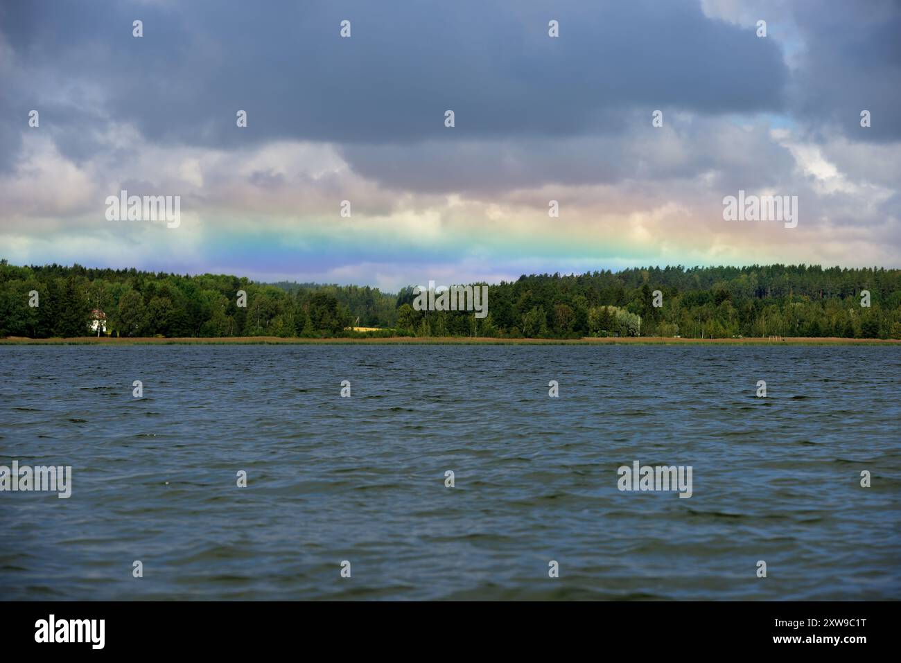 Questa immagine cattura un paesaggio naturale sereno e tranquillo caratterizzato da un lago calmo sotto un cielo coperto. Un arcobaleno sottile si estende attraverso l'orizzonte Foto Stock
