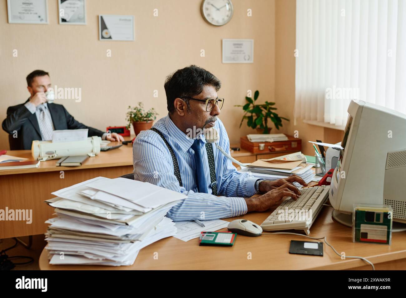 Uomo che indossa occhiali che parla al telefono e scrive al computer con le attrezzature dell'ufficio e i documenti sulla scrivania mentre il collega in background beve un caffè e osserva Foto Stock