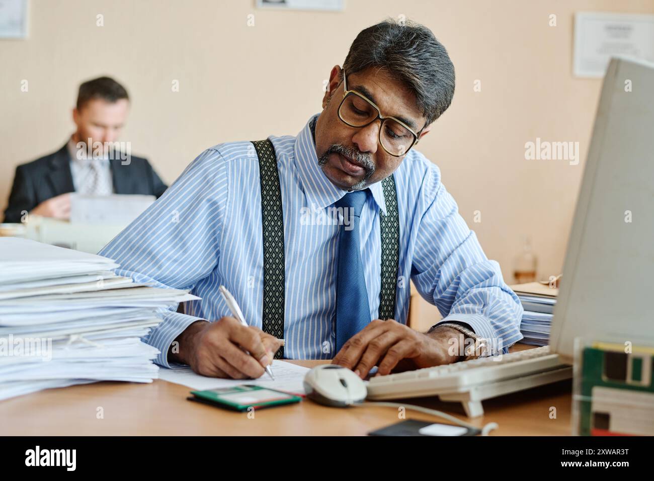Manager che esamina i documenti alla scrivania dell'ufficio, concentrandosi su un'espressione mirata. Pile di documenti e ambienti d'ufficio in background, a dimostrazione di un'atmosfera di lavoro intensa Foto Stock