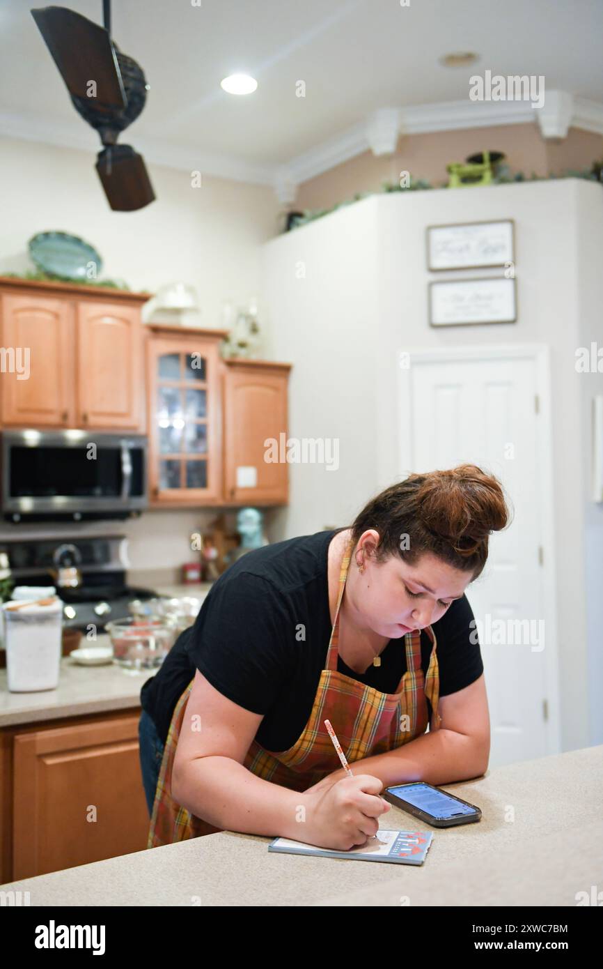 Donna che prende appunti in cucina con telefono e blocco note. Foto Stock